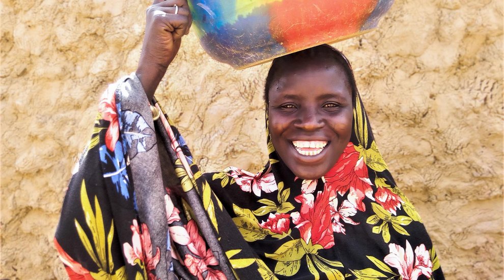 Eine burkinische Frau trägt eine Wasserschüssel auf dem Kopf und lacht in die Kamera